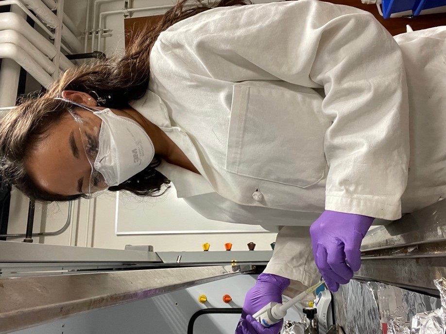 Image of Sarah working in the lab wearing a white robe and purple gloves holding the lab equipment